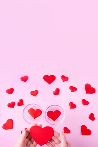 Hands hold a red heart shape, two glasses with hearts and white and red heart shapes on a pink background, top view - Foto, immagini