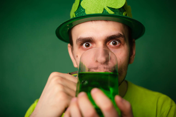 Muž v shamrockovém klobouku a zeleném tričku pije velký hrnek zeleného piva. Chlápek slaví den svatého Patricka sklenicí piva. - Fotografie, Obrázek