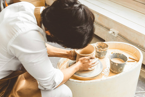 A young male potter is engaged in craft in his workshop on a potters wheel and makes a clay product - Foto, Bild