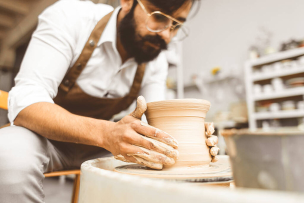 A young male potter is engaged in craft in his workshop on a potters wheel and makes a clay product - Zdjęcie, obraz