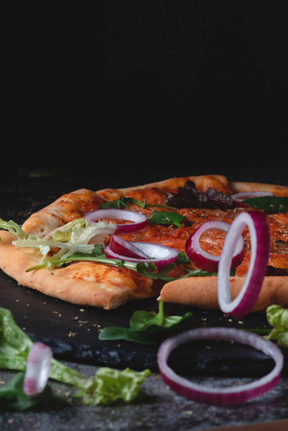 Hagyományos olasz pepperoni pizza hagymával, fűszernövényekkel, paradicsommal és sajttal sötét palatáblán és fekete háttérrel  - Fotó, kép