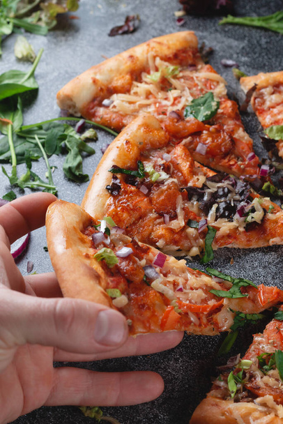 Kézzel veszi Pieces Hagyományos olasz pepperoni pizza paradicsommal, sajttal, hagymával és sok fűszernövényekkel, egy sötét palatáblán - Fotó, kép