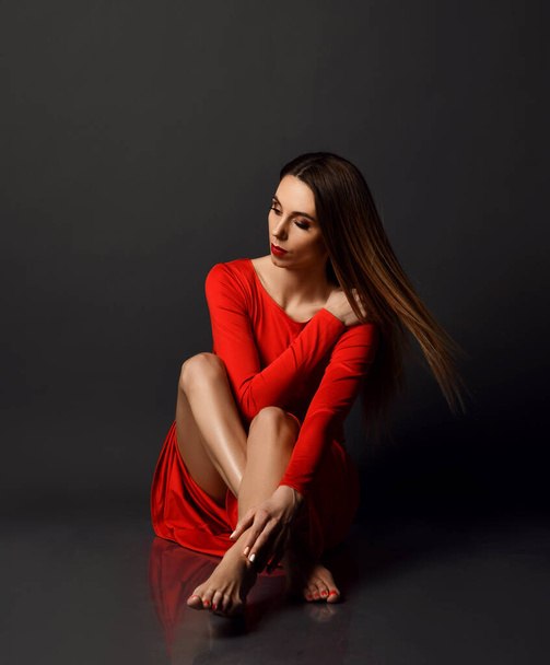 Sensual barefooted woman with long hair in red maxi dress sits on floor touching her leg and shoulder and looks down - Photo, image