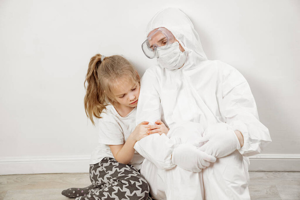 Pieni tyttö halailee lääkäriä valkoisessa suojapuvussa, naamiossa, laseissa ja käsineissä. - Valokuva, kuva