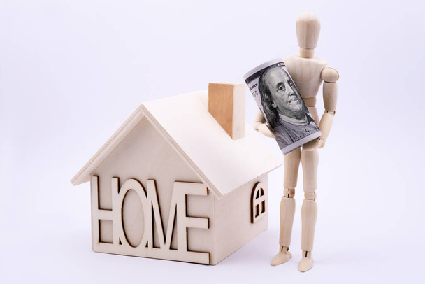 Izolovaný dřevěný jednoduchý domeček na hraní a dřevěná figurka symbolické osoby na bílém pozadí držící stodolarovou bankovku - Fotografie, Obrázek