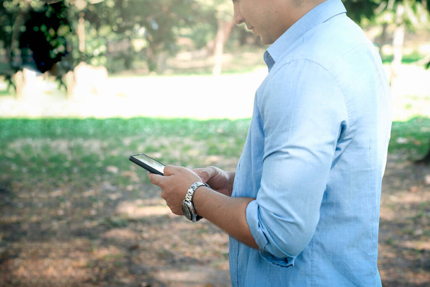 Młody przystojny mężczyzna w luźnej sukience, korzystający ze smartfona w ogrodzie, oglądający telefony komórkowe i czytający wiadomości  - Zdjęcie, obraz