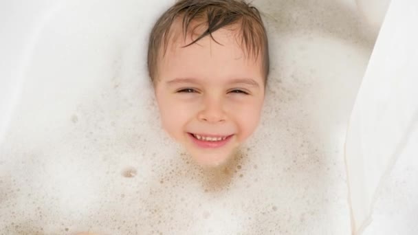 Portrait of cheerful little boy lying in bath with soap foam and looking in camera. Concept of child hygiene and health care at home. Child having fun and playing at home. - Footage, Video