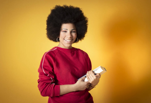 mujer afro bonita, con ropa casual, con un libro, sonriendo feliz, fondo amarillo, espacio para copiar - Foto, Imagen