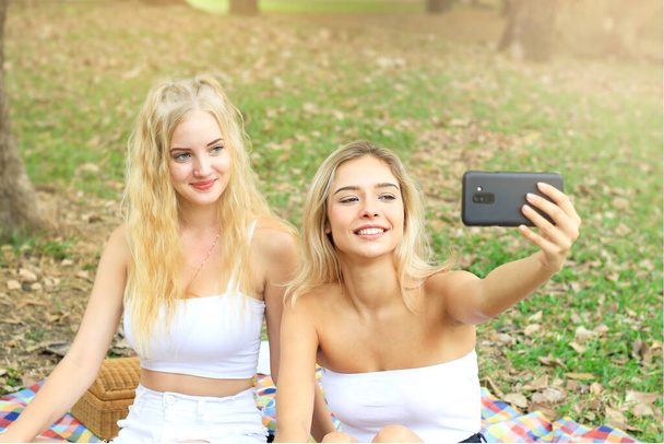 公園でピクニックをしている幸せな友人、 2人の若い美しい10代の女の子がスマートフォンで自撮りし、夏の庭で一緒に時間を過ごしながら楽しい時間を過ごしています、女性の屋外友情の概念 - 写真・画像