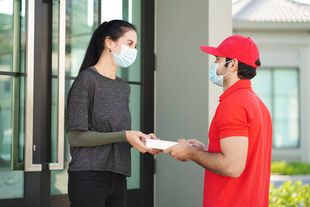 Beautiful customer wearing a surgical mask receives a food box from a food delivery man, wears a red uniform, and wears a mask delivered to the apartment. New normal concept.  - Valokuva, kuva