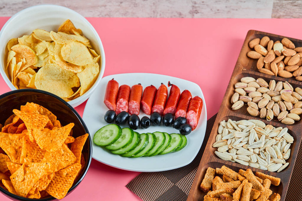 Разные закуски на розовом столе. Чипсы, сосиски, фисташки, оливки - Фото, изображение