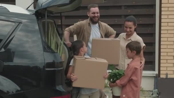  Portrét šťastných rodičů a malých dětí držících rostliny a lepenkové krabice a pózujících vedle svého auta zaparkovaného před jejich novým domem - Záběry, video