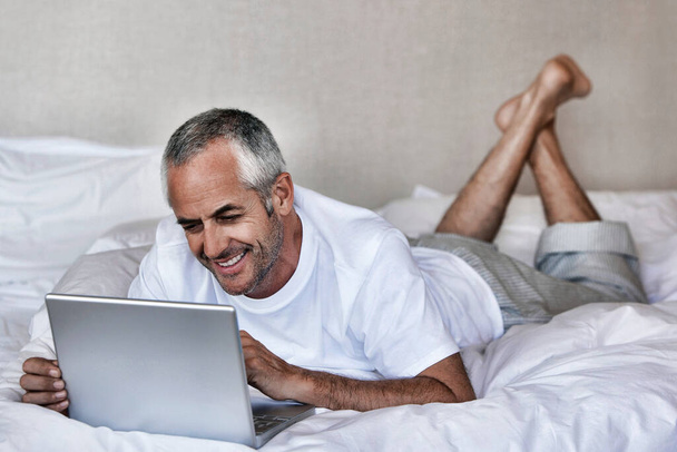 Happy middle aged man using laptop while lying in bed at home - Valokuva, kuva