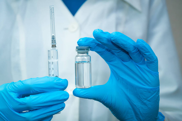 Kobieta w białym płaszczu, niebieskie rękawiczki trzymają strzykawkę w jednej ręce, a w drugiej butelkę ze szczepionką, zbliżenie leku. Koncepcja leku, leków i szczepień - Zdjęcie, obraz