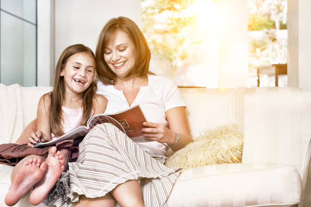 Alegre madre e hija leyendo libro en la sala de estar durante el encierro - Foto, imagen
