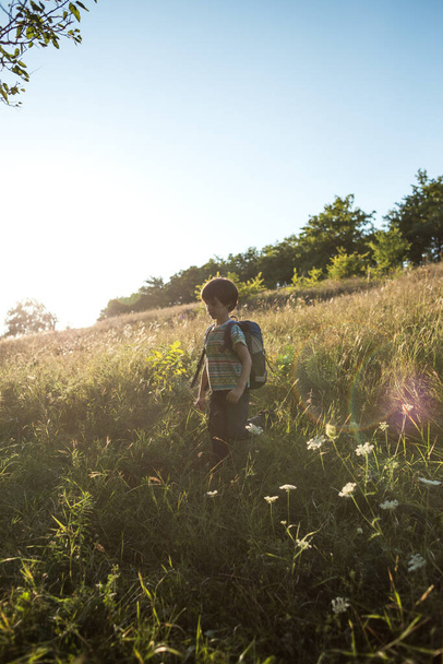 Een jongen met een rugzak wandelt door de weide, een kind verkent de natuur, een kind staat alleen tussen de bomen, een portret van een jongen. - Foto, afbeelding