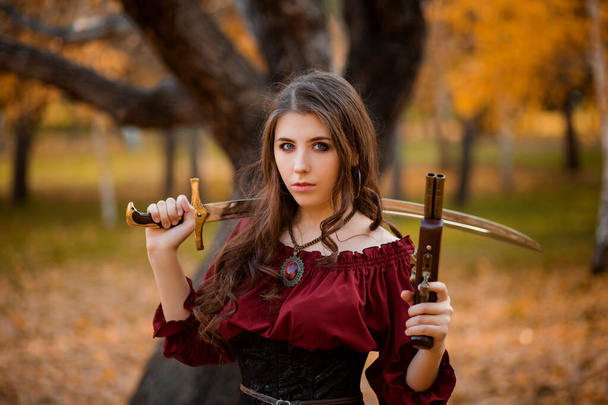 Portrait of a beautiful, young woman with a saber and a pistol in her hands against the backdrop of a large tree and an autumn park. - Foto, immagini