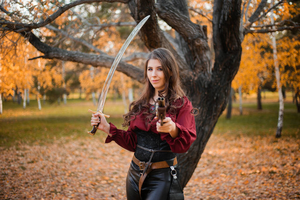 A beautiful girl in a shirt, corset and tight trousers with a saber and a pistol in her hands against the background of an autumn park in the evening. Adventurer woman concept. - Фото, зображення