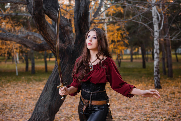 A young woman holds a saber while standing against the backdrop of a gloomy, dark autumn park. - Photo, Image