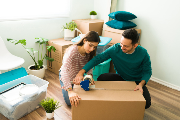 Happy boyfriend and girlfriend packing and putting tape on a cardboard box. Young woman and hispanic man moving out of their home or apartment - Photo, Image