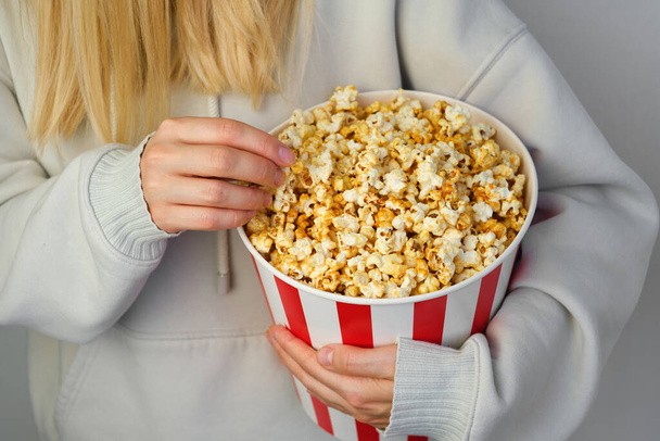 Popcorn paper bucket in the hands of a young girl preparing to watch a movie. Showtime. Eating delicious unhealthy sweet snacks. Going to cinema for a new film. Rest and entertainment. Popcorn closeup - Photo, image