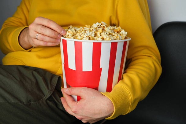 Popcorn paper bucket in the hands of a men preparing to watch a movie. Showtime. Eating delicious unhealthy sweet snacks. Going to cinema for a new film. Rest and entertainment. Popcorn closeup - Foto, Bild