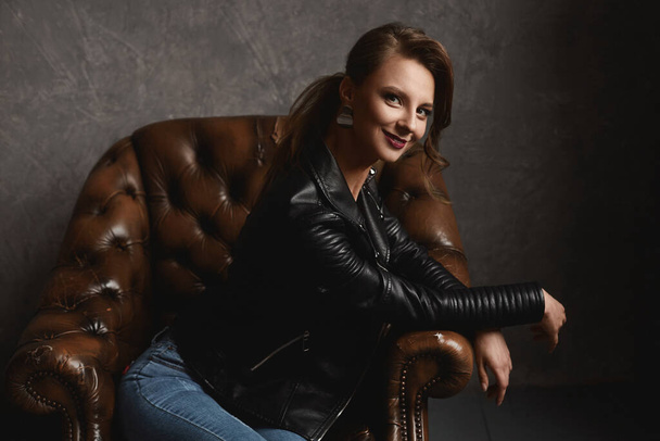 Slim model girl with perfect makeup in stylish leather jacket and jeans sits in a vintage armchair near the grey wall - Photo, image