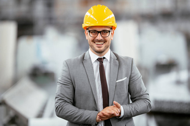 Retrato de hombre de negocios en un traje gris con un casco protector amarillo en fábrica. - Foto, Imagen