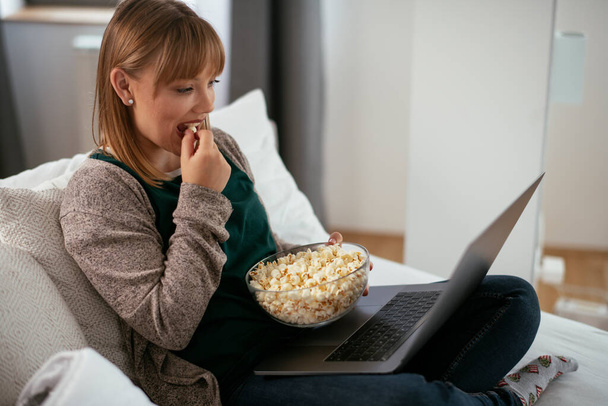 Güzel sarışın kadın dizüstü bilgisayarda film izliyor ve patlamış mısır yiyor. - Fotoğraf, Görsel