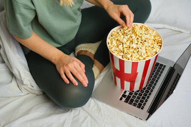 Popcorn bucket in the hands of a young girl preparing to watch a movie in bed on the laptop. Showtime. Eating delicious sweet snacks. Watching new film in home. Rest and entertainment in bedroom - Fotografie, Obrázek