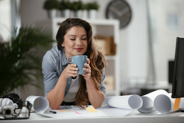 Молода випадкова жінка п'є каву в офісі. Приваблива жінка-менеджер сидить за столом після важкого робочого дня
 - Фото, зображення