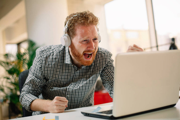 Businessman in office. Handsome ginger man listening music in headphones - Photo, Image