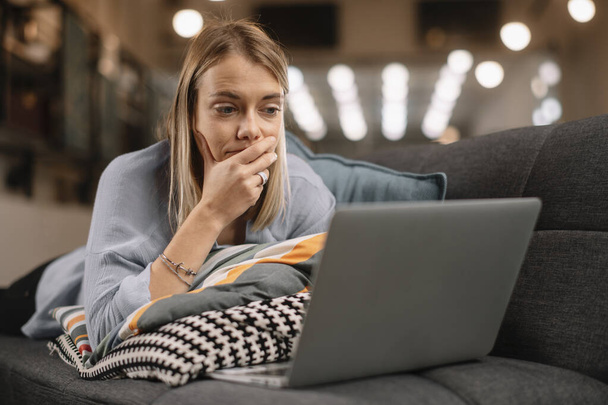 Молодая женщина лежит на диване и смотрит фильм на ноутбуке - Фото, изображение