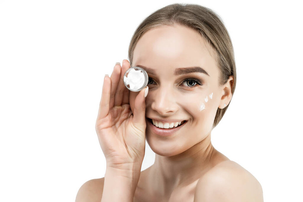 Close up of an attractive woman with fresh and well-groomed skin, cream on her face and she is holding a box of cream, she is sincerely smiling at the camera. - Photo, image