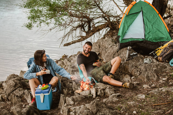  Pár szerelmes kempingező erdei túrára. Kaja túrázáshoz és kempingezéshez. A pár a tábortűz mellett ül és eszik és iszik.. - Fotó, kép