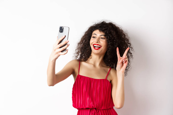 Елегантна молода жінка в червоній сукні бере селфі на смартфон, позує на вечірці, стоїть з мобільним телефоном на білому тлі
 - Фото, зображення