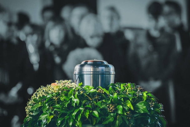 Een metalen urn met as van een dode op een begrafenis, met rouwende mensen op de achtergrond op een herdenkingsdienst. Droevig treurmoment aan het einde van een leven. Laatste afscheid van een persoon in een urn. - Foto, afbeelding