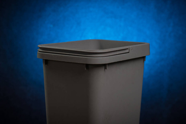 Серый пластиковый мусорный бак с открытой верхней и видимой ручкой на растворяющемся синем фоне в студии. Концептуальная фотография пустой пластиковой современной мусорной корзины для разделения мусора. - Фото, изображение