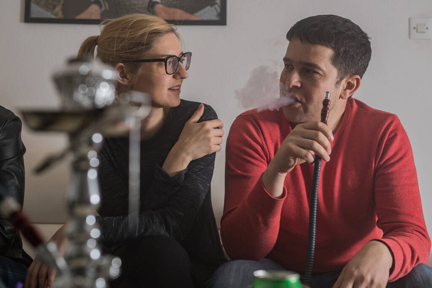 Jovem loira europeia com e um homem de camisola vermelha fumando uma shisha em uma sala de estar em casa. Fumaça de lazer em uma festa em casa. Shisha é visível no primeiro plano borrado. - Foto, Imagem