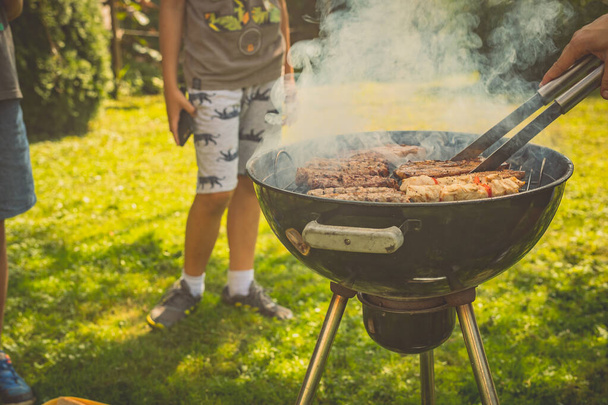 Kinderen wachten om vlees te ontvangen van een grill terwijl ze er achter wachten. Ronde grill in rook met cevapcici op een tuin gazon met een hand draaien vlees. - Foto, afbeelding