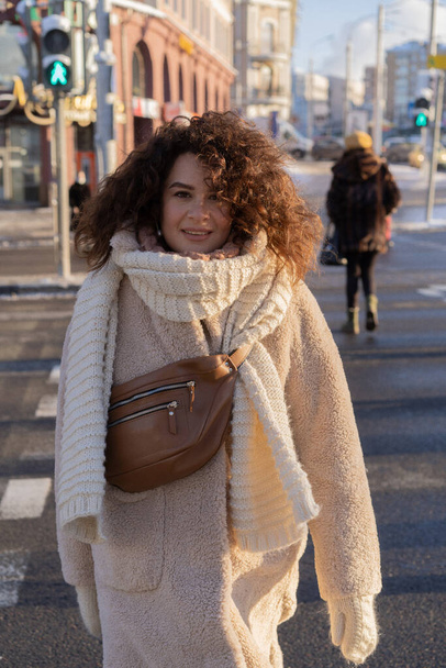 Young woman with dark curly hair, close-up portrait, warmly dressed scarf, winter frost, sunny day on the street in the city. - Фото, изображение