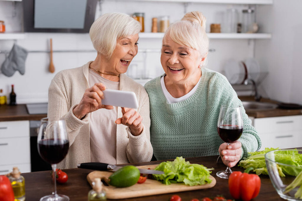 весела старша жінка вказує пальцем на смартфон поруч з другом і овочами на столі
 - Фото, зображення