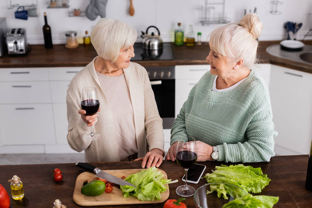 赤ワインと一緒にグラスを持ってテーブルの上で友人や野菜を見ている陽気な先輩女性の高い角度のビュー - 写真・画像