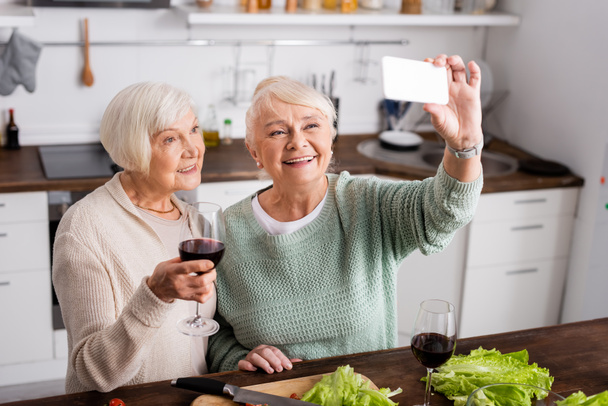 alegre anciana sosteniendo copa con vino tinto y mirando el teléfono inteligente mientras su amigo se toma selfie en la cocina  - Foto, Imagen