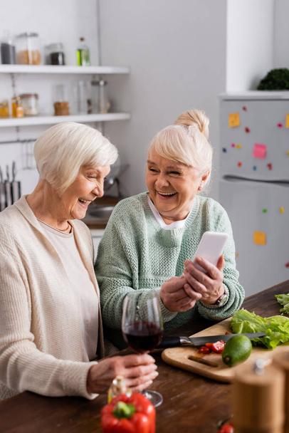 iloinen vanhempi nainen tilalla älypuhelin lähellä eläkkeellä ystävä keittiössä  - Valokuva, kuva