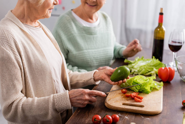cropped view of senior woman holding knife and avocado near retired friend in kitchen  - Foto, afbeelding