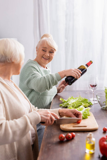 happy senior woman pouring red wine in glass and looking at retired friend cutting cherry tomato on blurred foreground - 写真・画像
