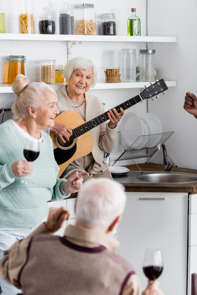 幸せな引退した女性は台所で高齢者の異人種間の友人の近くでアコースティックギターを演奏  - 写真・画像