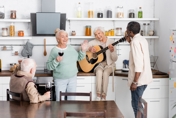 onnellinen vanhempi nainen soittaa akustista kitaraa lähellä iloinen monikulttuurinen ystäviä lasit viiniä keittiössä  - Valokuva, kuva