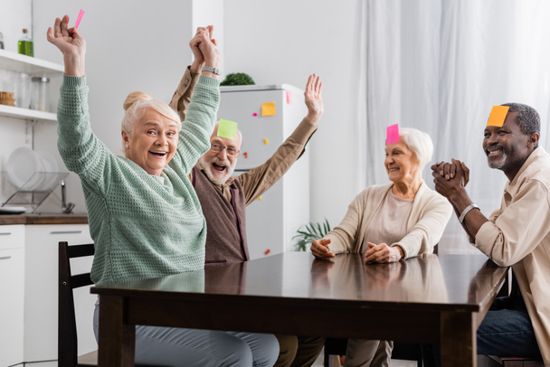 excited and multicultural senior friends with sticky notes on foreheads playing game in kitchen  - Photo, Image